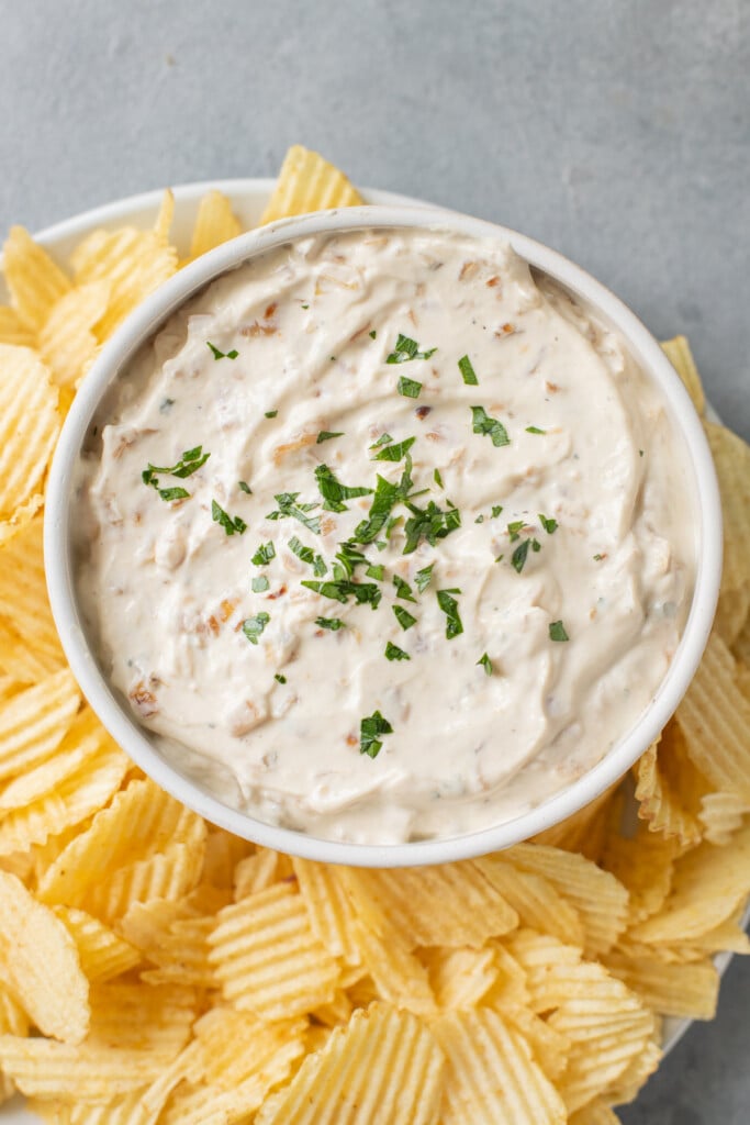 An overhead shot of a bowl of Healthy Onion Dip with potato chips on the side on a grey background