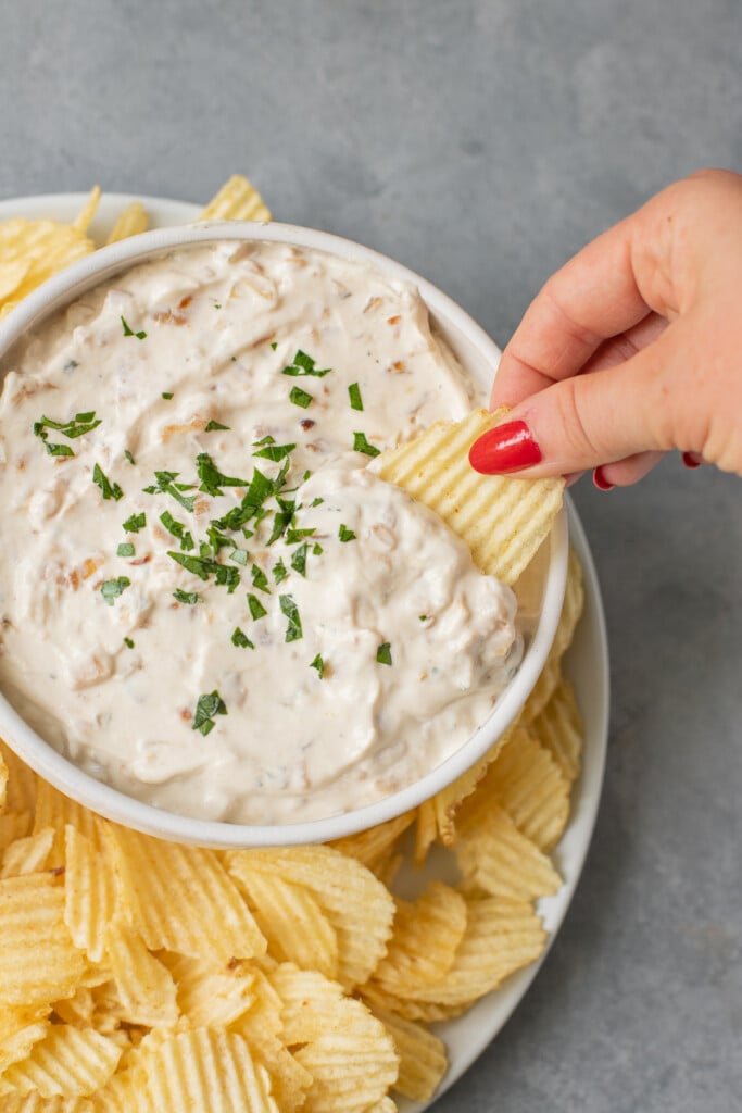 A bowl of french onion dip made with caramelized onions with a hand dipping in a chip with chips on the side on a grey background