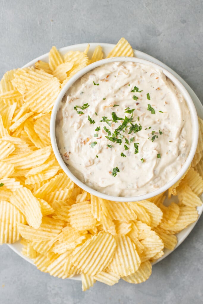 An overhead shot of a big plate of potato chips and a bowl of Healthy Onion Dip on a grey background