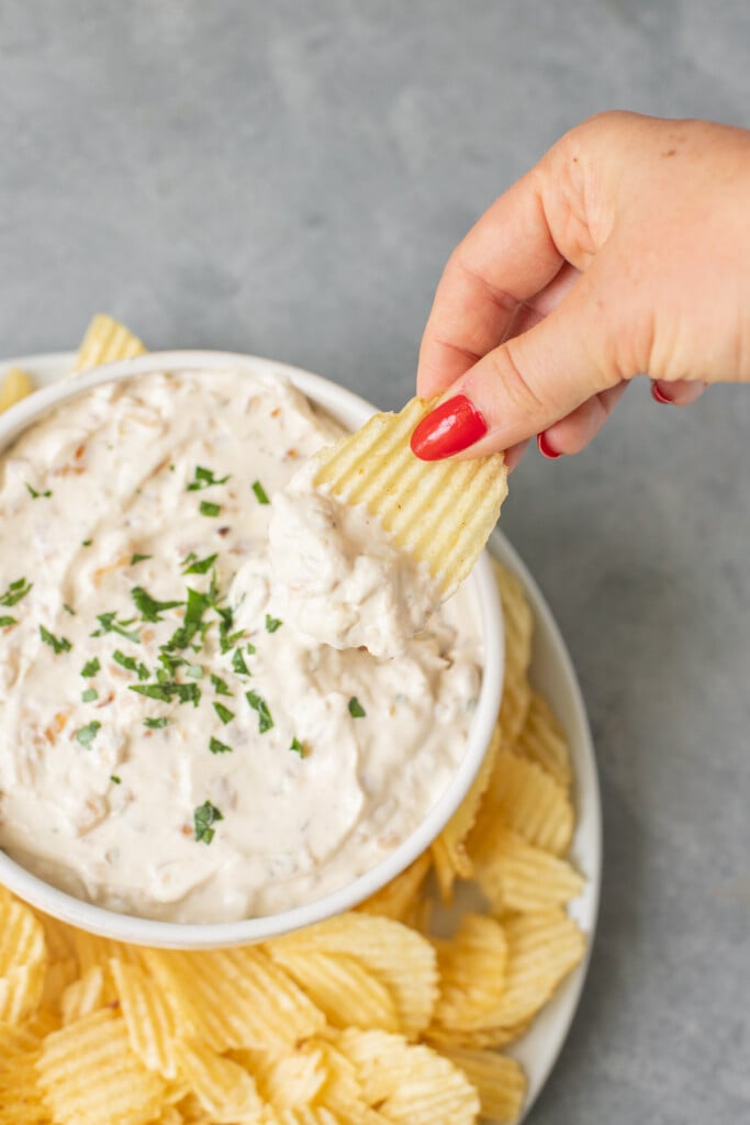 A bowl of french onion dip with a hand dipping in a chip with chips on the side on a grey background