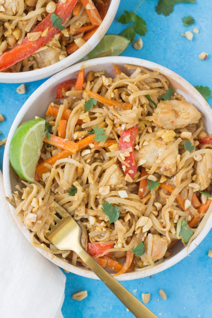 An overhead views of Healthy Pad Thai on a bright blue background. There's a gold fork in the bowl with a swirl of healthy noodles waiting to be eaten.