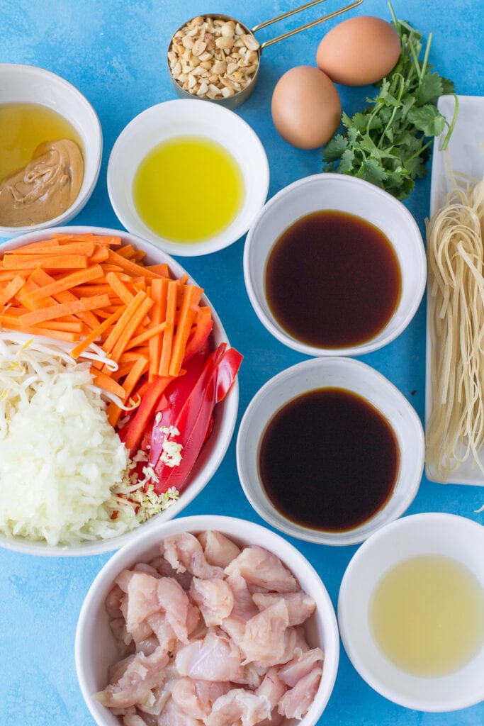 Ingredients for Healthy Pad Thai in small white bowls on a bright blue background - veggies, honey, oil, fish sauce, coconut aminos, rice wine vinegars, peanuts, noodles and chicken.