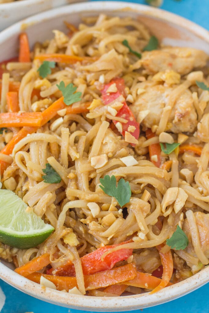 A close up of a white bowl of pad thai noodles on a bright blue background. Peppers, carrots and bean sprouts are tossed with the saucy noodles with crushed peanuts on top.