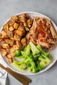 Healthy Crockpot Pulled Pork in a white dish with roasted potatoes and broccoli off to the side