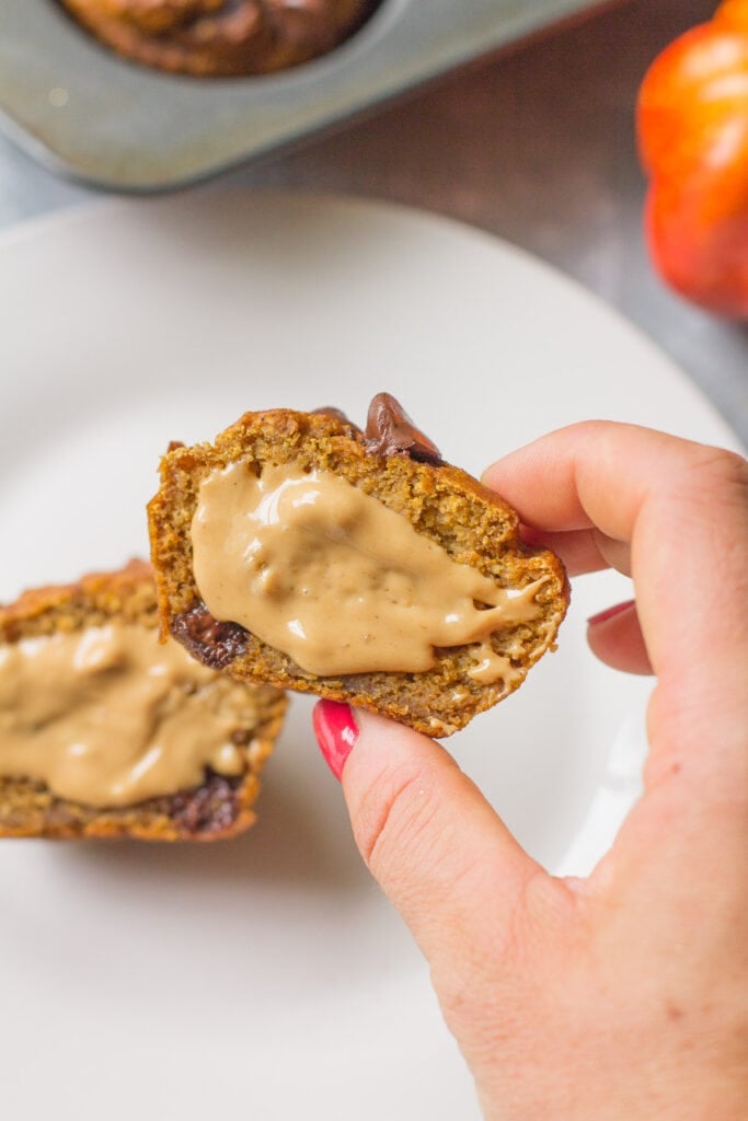 A healthy pumpkin muffin cut in half with nut butter spread on top. Muffin tin and small pumpkin are in the background behind the plate.