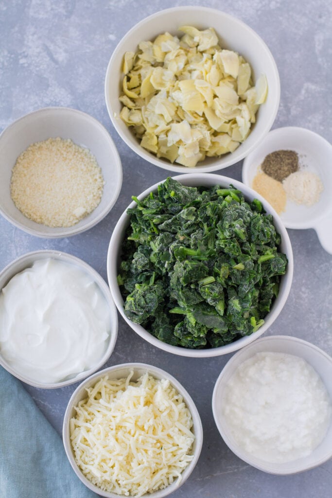 ingredients for healthy spinach artichoke dip laid out in small white bowls on a grey background. There is no cream cheese or mayo in this recipe.