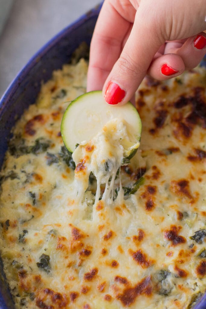 A zucchini chip scooping up healthy spinach artichoke dip from a baking dish.