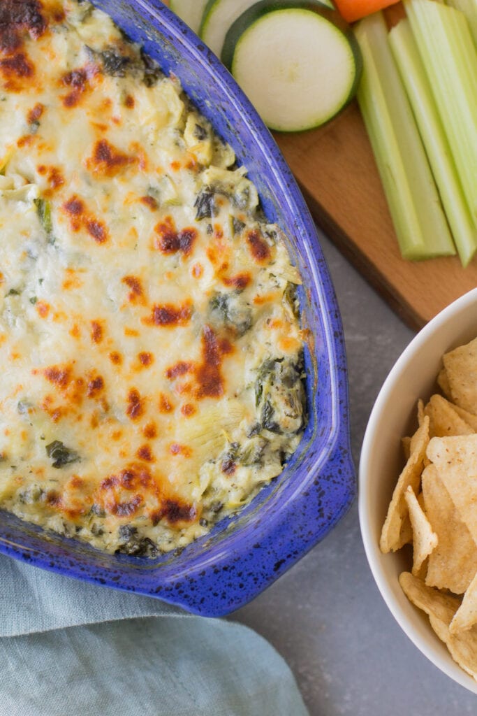 A close up of healthy spinach artichoke dip on a grey background with chips and veggies surrounding it.