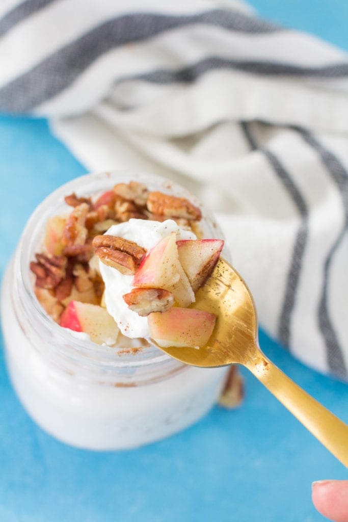 an apple pie healthy yogurt parfait with a gold spoon taking a scoop out of the jar + holding it up to the camera.