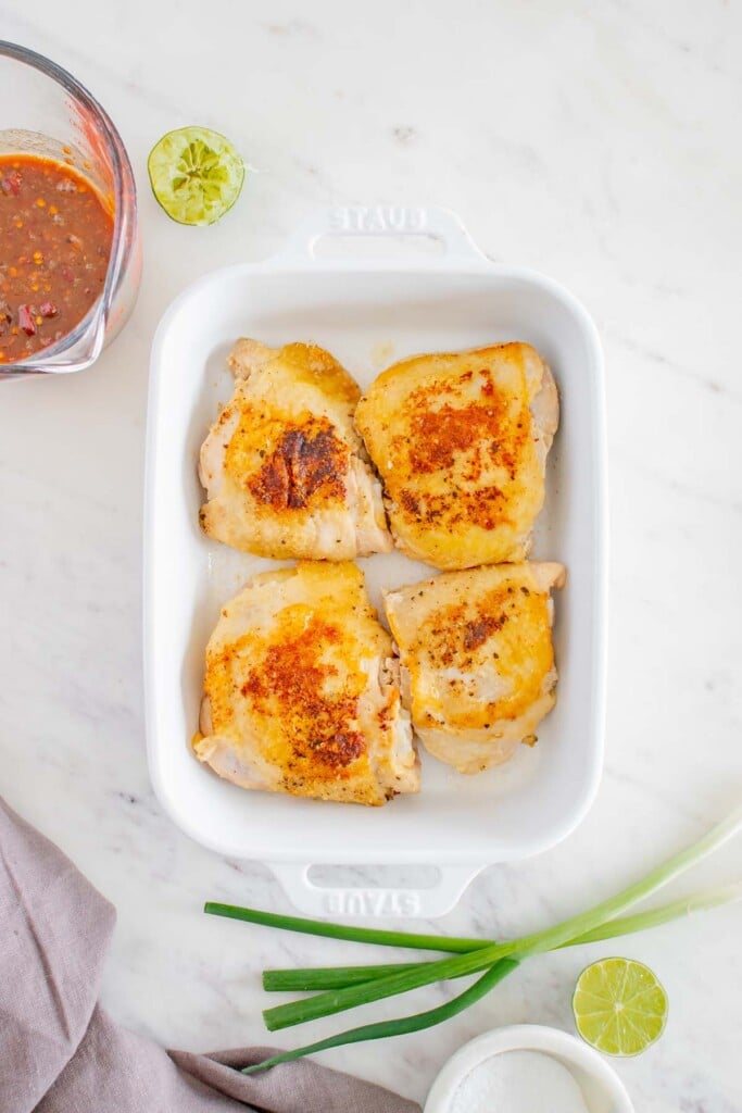 4 browned chicken thighs in a white baking dish with a honey chipotle sauce in a clear measuring cup on the side on a white background
