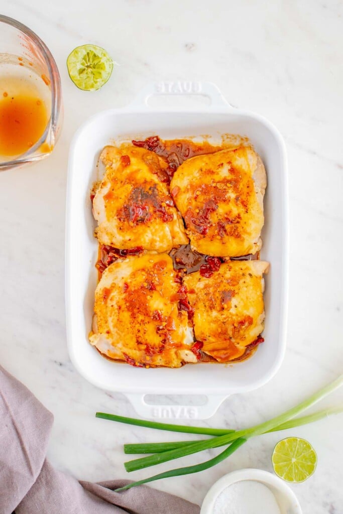4 browned chicken thighs covered in a honey chipotle sauce in a white baking dish with a clear measuring cup on the side on a white background