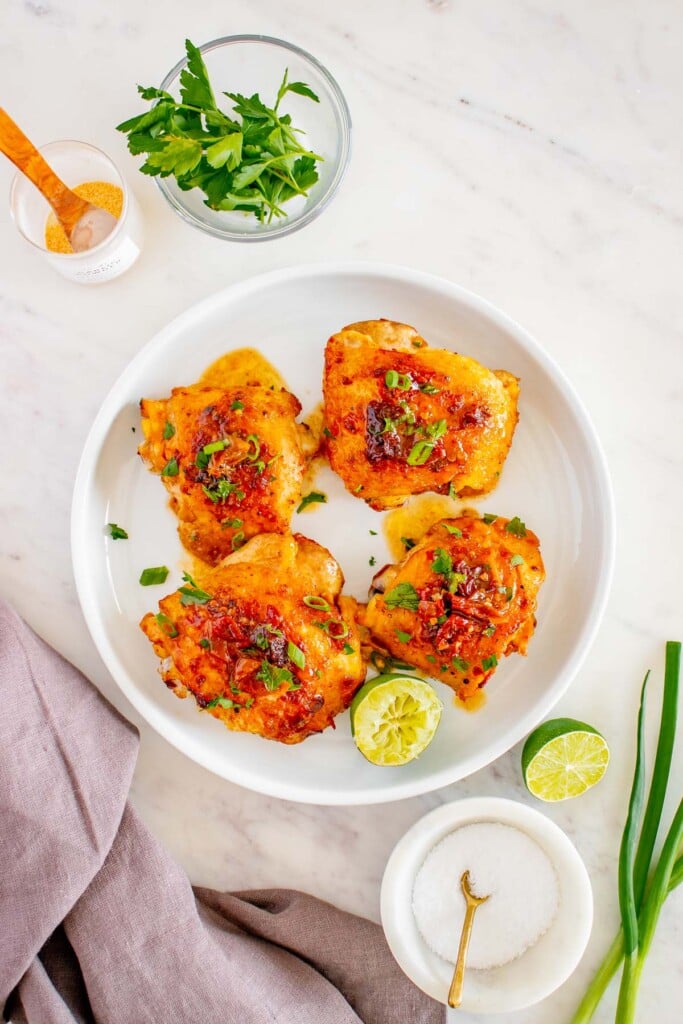 An overhead shot of 4 honey chipotle chicken thighs on a white plate with fresh parsley and green onions on the side on a white background