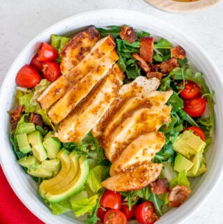 An overhead shot of a white bowl of Honey Mustard Chicken Salad with tomatoes, avocado and bacon on a bed of mixed greens