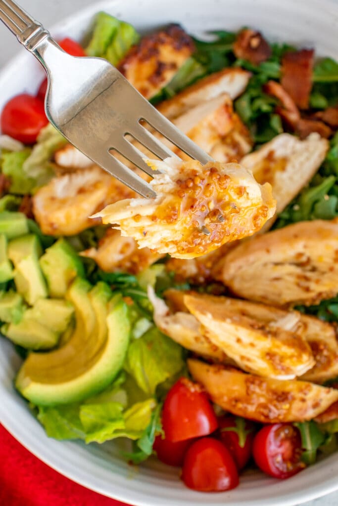 A fork with a piece of honey mustard chicken with a salad in the background with more chicken, bacon, tomatoes, avocado on a bed of mixed greens