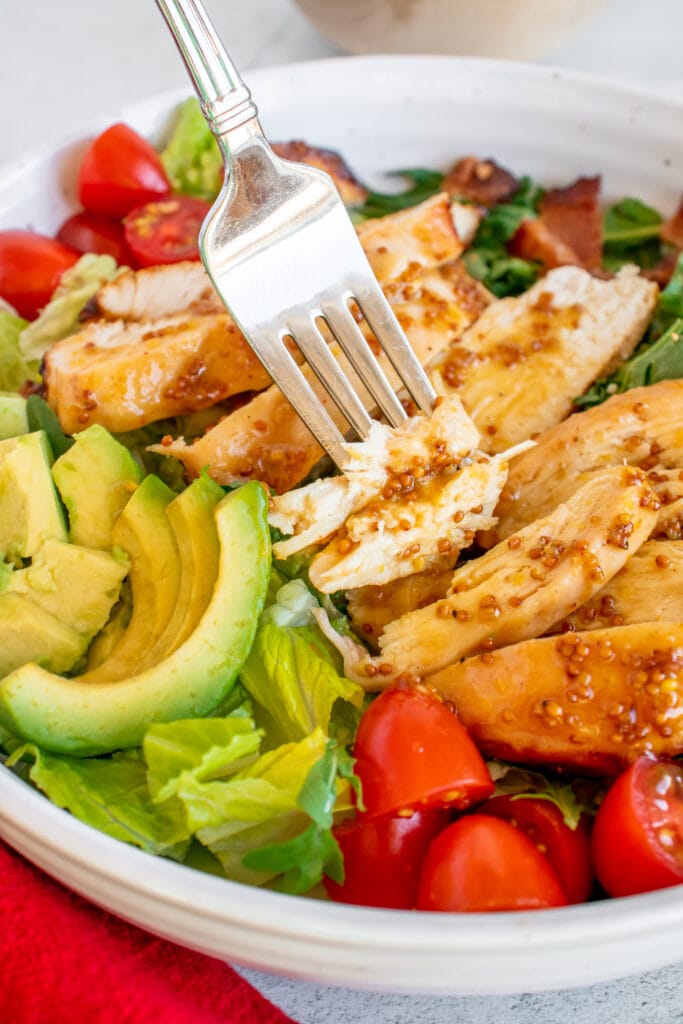 A fork taking a piece of chicken from a bowl of Honey Mustard Chicken Salad with avocado and tomatoes on it