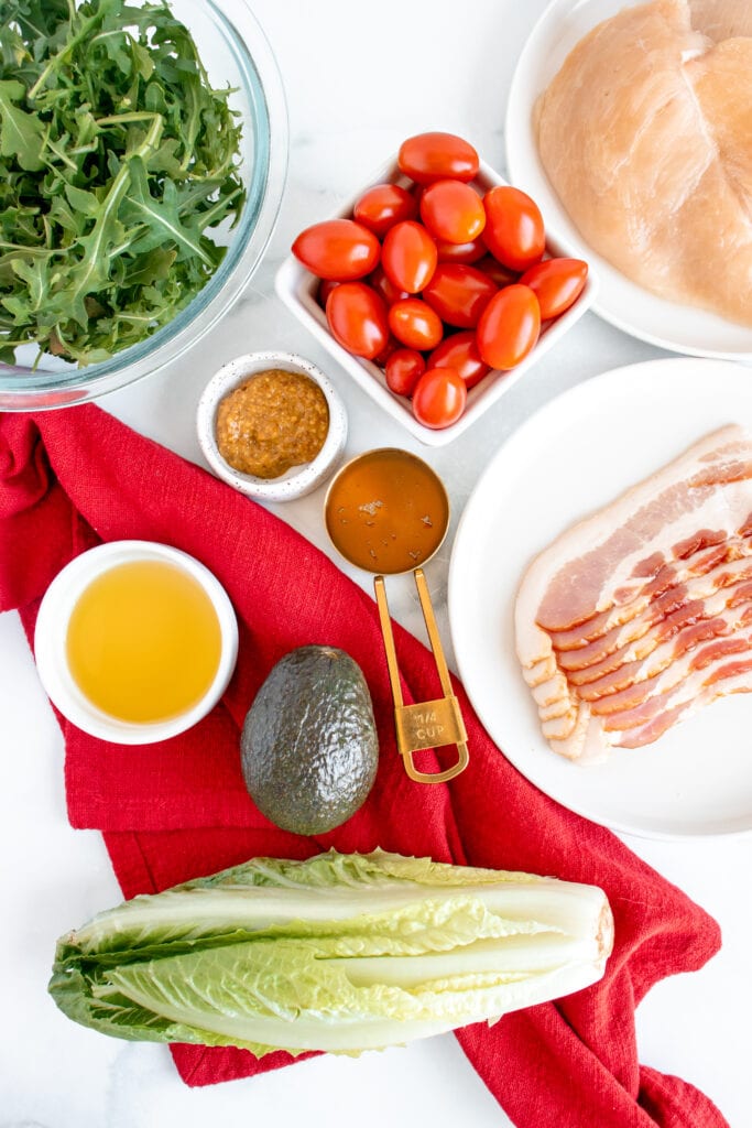 Ingredients for Honey Mustard Chicken Salad in white bowls: chicken breasts, bacon, romaine lettuce, arugula, cherry tomatoes, avocado, honey, whole grain mustard, white wine vinegar, olive oil, salt, pepper