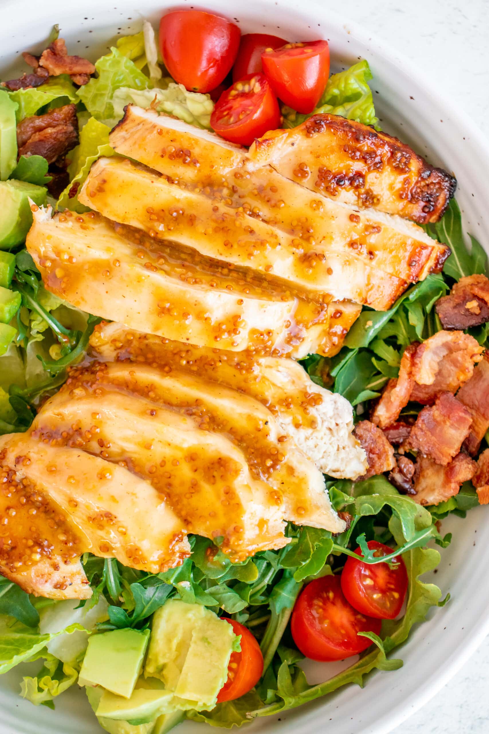 A white bowl of Honey Mustard Chicken Salad with tomatoes, avocado and bacon on a bed of mixed greens