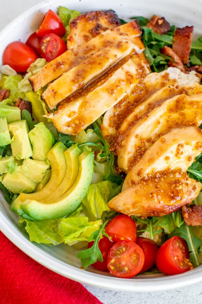 A white bowl of Honey Mustard Chicken Salad with tomatoes, avocado and bacon on a bed of mixed greens