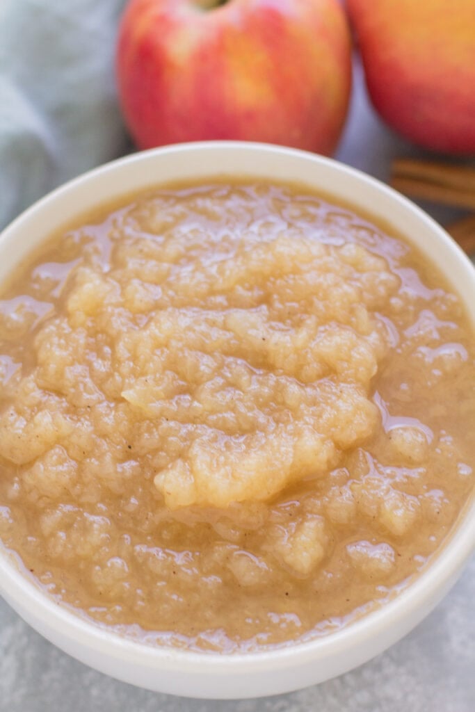 Instant pot applesauce in a white bowl on a blue background with apples and spices in the background.