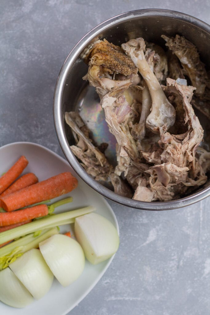 Ingredients for instant pot bone broth on a grey background. a plate with veggies is off to the left and a pot with chicken bones is off to the right