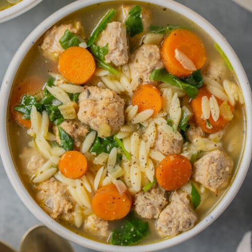An overhead shot of Italian wedding soup in a white bowl with gold spoons and a bowl on the side on a grey background