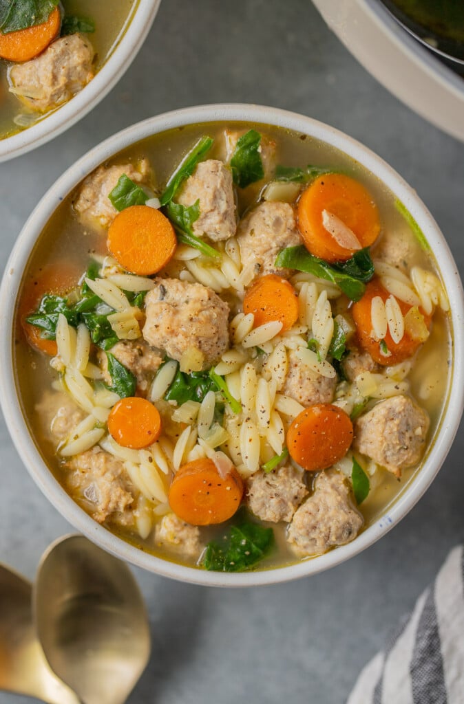 An overhead shot of Italian wedding soup in a white bowl with gold spoons and a bowl on the side on a grey background