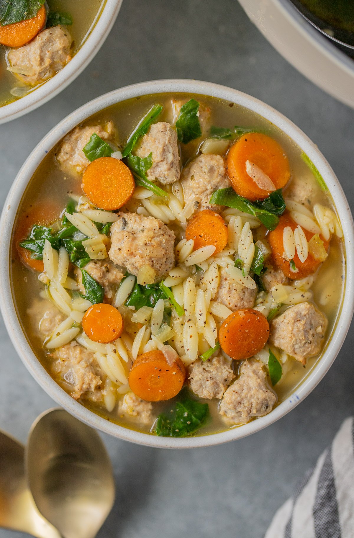 An overhead shot of Italian wedding soup in a white bowl with gold spoons and a bowl on the side on a grey background