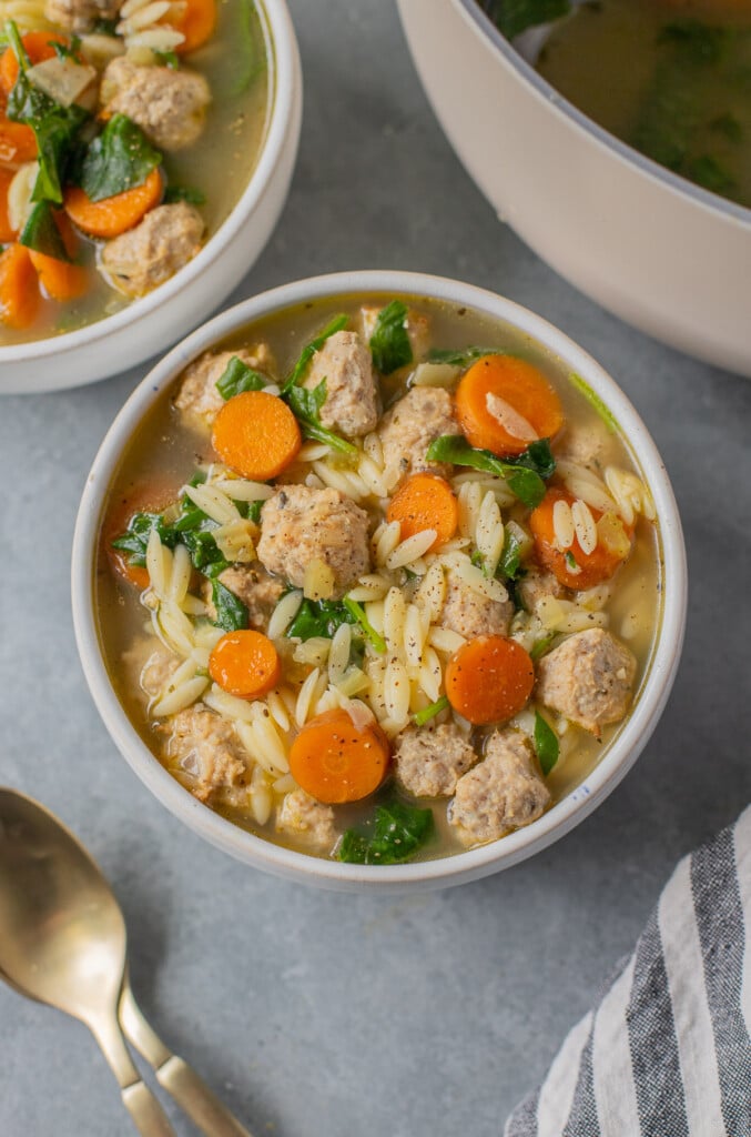 An overhead shot of Italian wedding soup in a white bowl with gold spoons and a bowl of soup on the side on a grey background