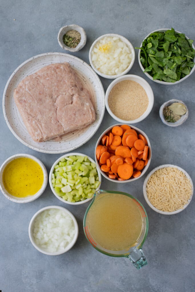 Ingredients for Italian Wedding Soup in white bowls: ground turkey, egg, breadcrumbs, olive oil, garlic, onion, celery, carrots, rosemary, parsley, basil, pepper, bay leaves, chicken broth or beef broth, fresh spinach, orzo pasta