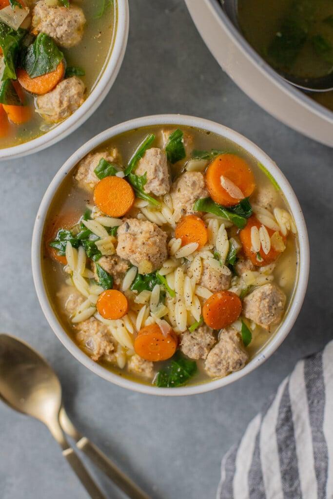 An overhead shot of Italian wedding soup in a white bowl with gold spoons and a bowl of soup on the side on a grey background