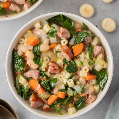 An overhead shot of a bowl of kielbasa soup with another bowl of soup and spoons on the side