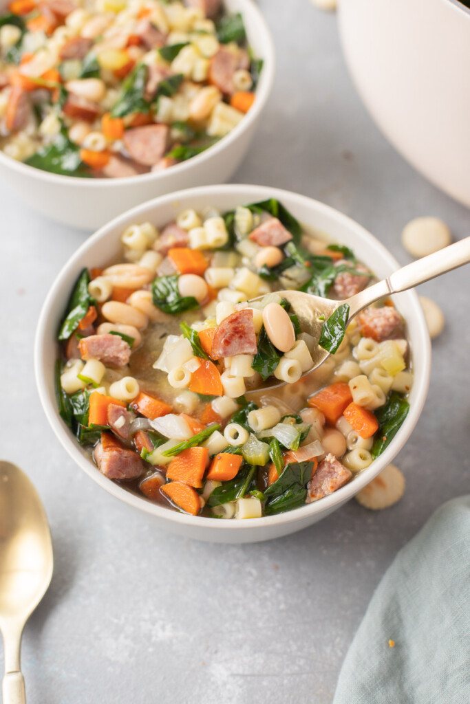 A bowl of polish sausage soup with a spoon taking a bite out of it with another bowl of soup in the background 