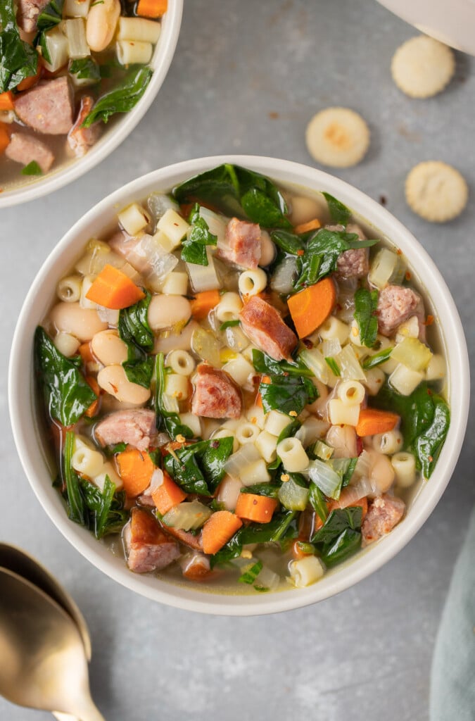 An overhead shot of a bowl of kielbasa soup with another bowl of soup and spoons on the side on a grey background
