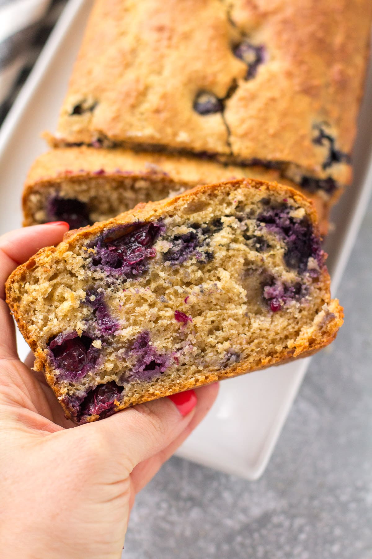 A hand holding up a slice of Lemon Blueberry Bread off a white plate