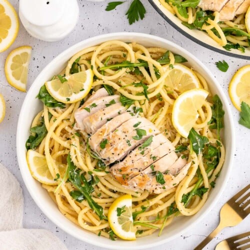 Lemon Chicken Pasta in a white bowl garnished with fresh parsley and lemon slices with a dutch oven and forks on the side