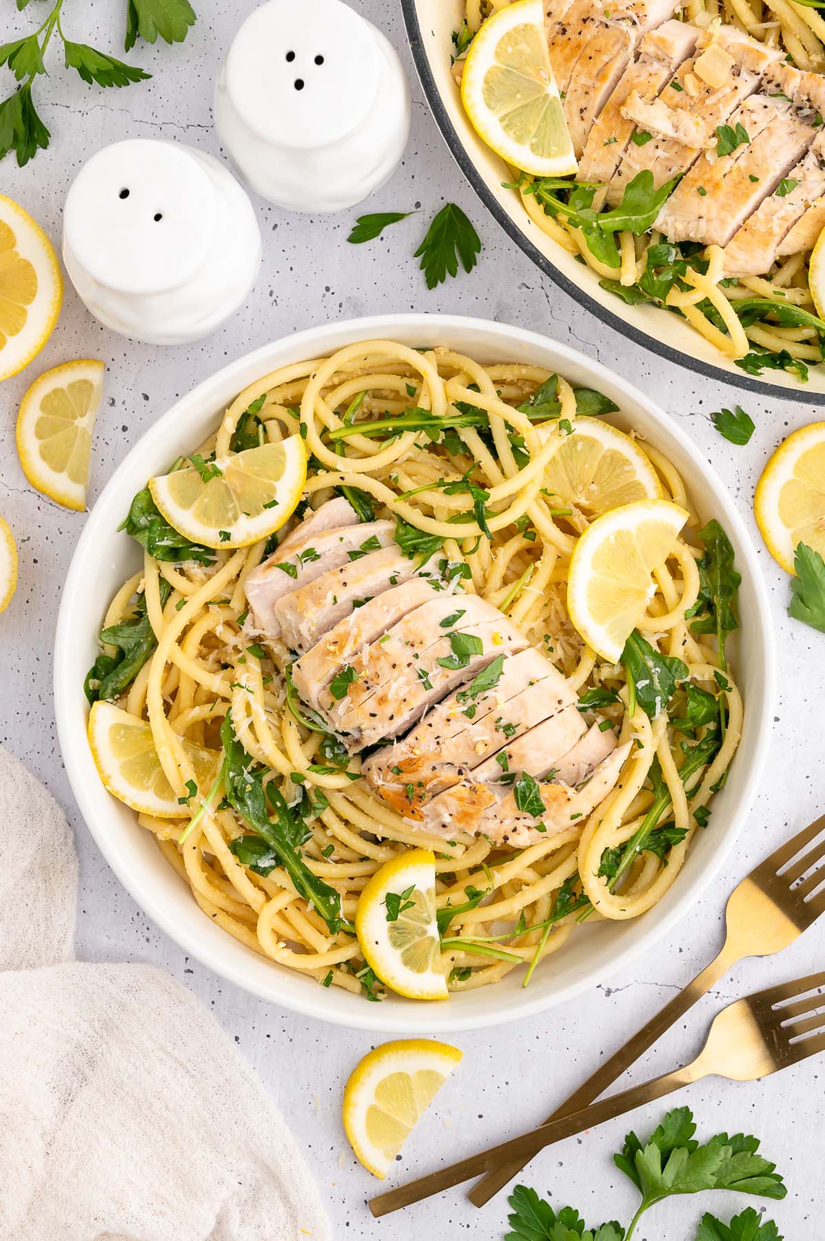 Lemon Chicken Pasta in a white bowl garnished with fresh parsley and lemon slices with a dutch oven and forks on the side