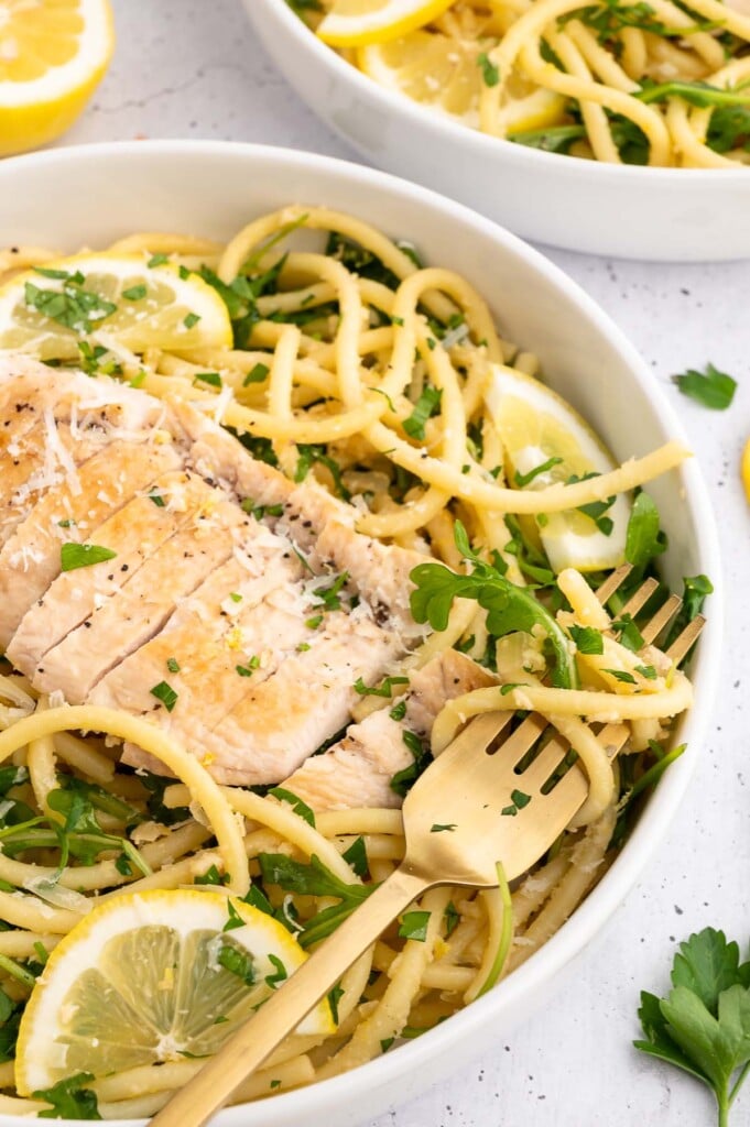 A fork spun in pasta in a white bowl of lemon chicken pasta garnished with fresh parsley and lemon slices