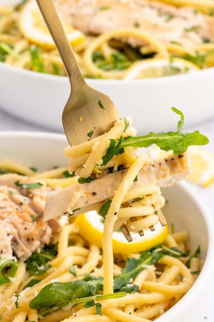 A fork holding up a bite of lemon chicken pasta with a white bowl of the chicken pasta with lemon sauce in the background