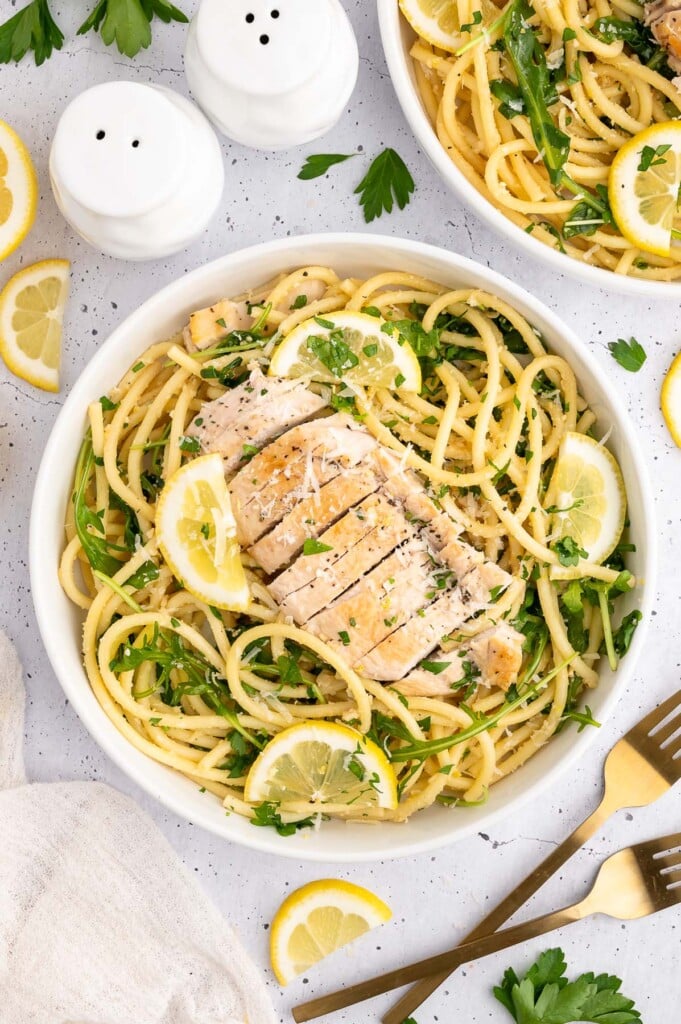 Lemon Chicken Pasta in a white bowl garnished with fresh parsley and lemon slices with a dutch oven and forks on the side
