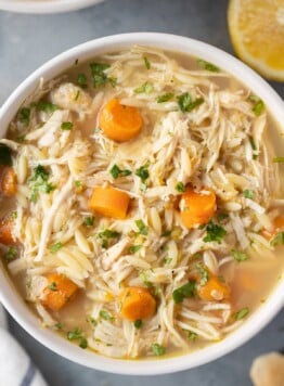 A close up of Lemon Chicken Soup in a white bowl on a grey background with lemons and soup off to the side