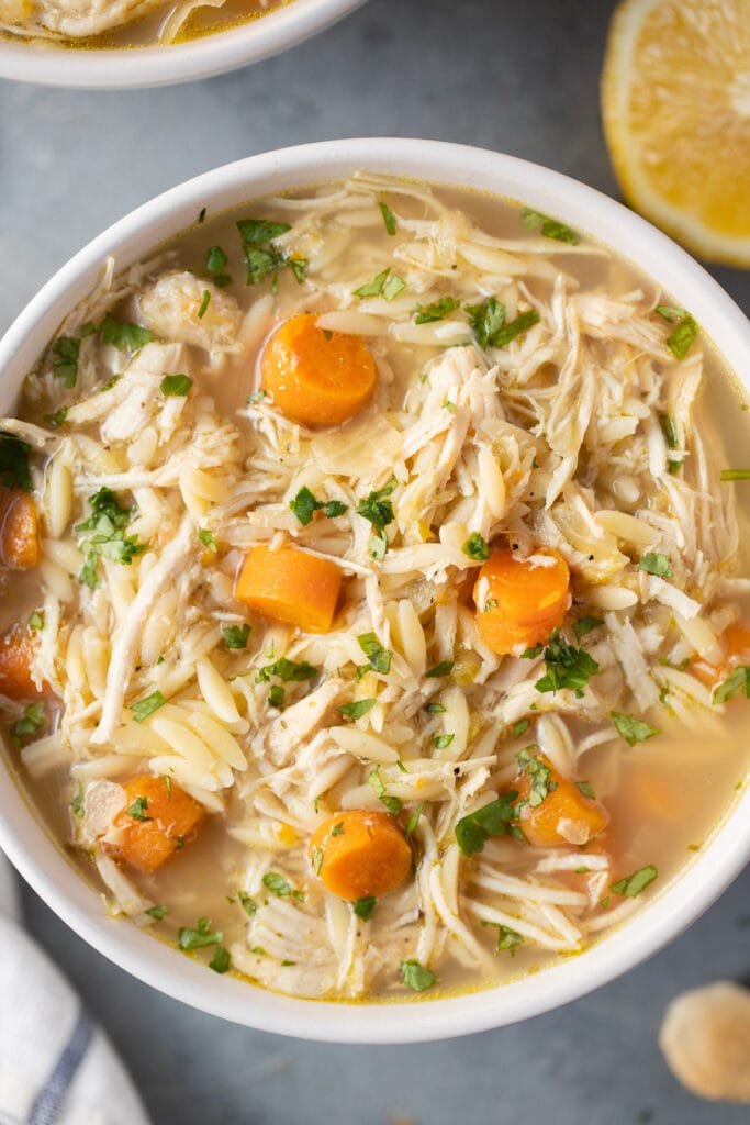 A close up of Lemon Chicken Soup in a white bowl on a grey background with lemons and soup off to the side