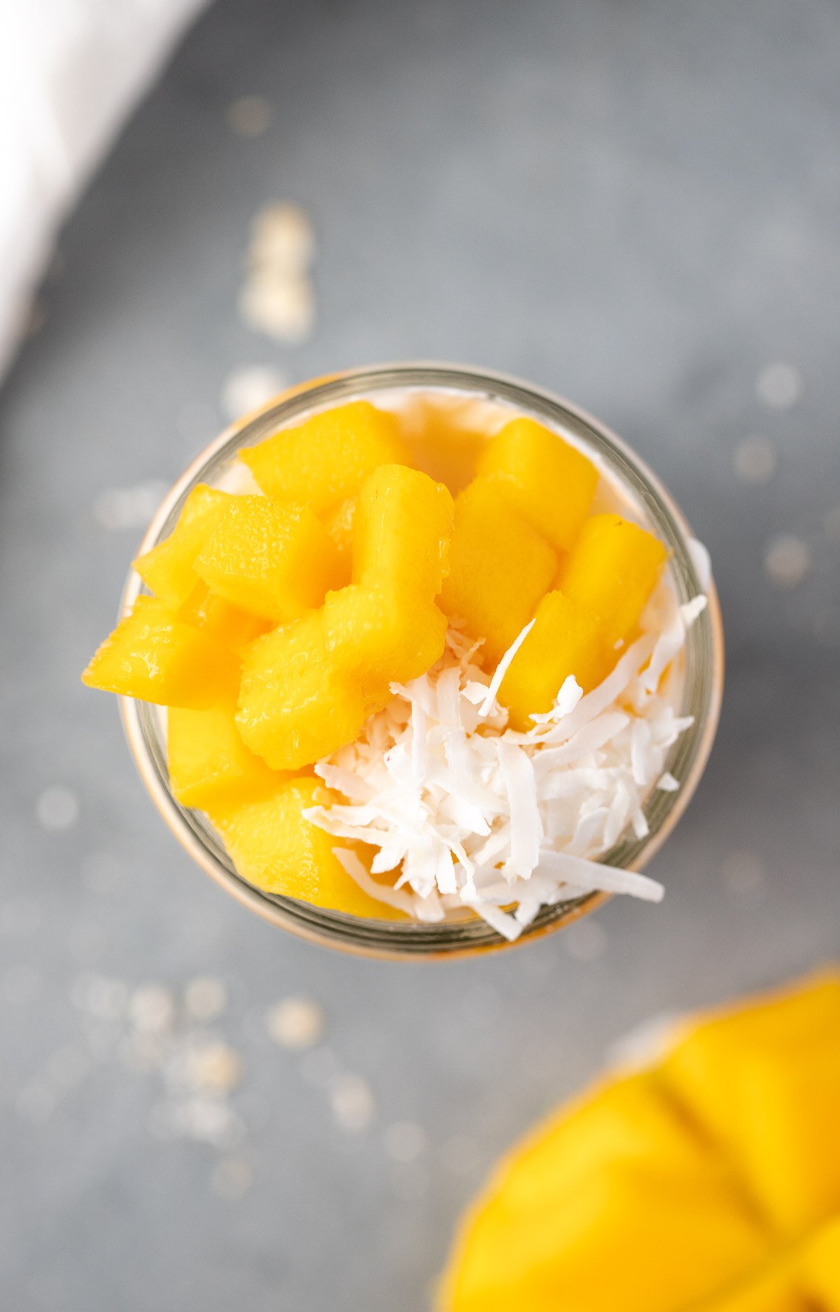 An overhead shot of mango overnight oats in a mason jar with mango in the background on a grey background