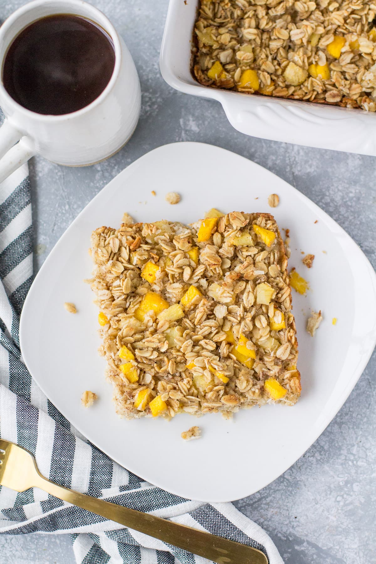 An overhead shot of mango pineapple oatmeal on a white plate with coffee and a baking dish off to the side.