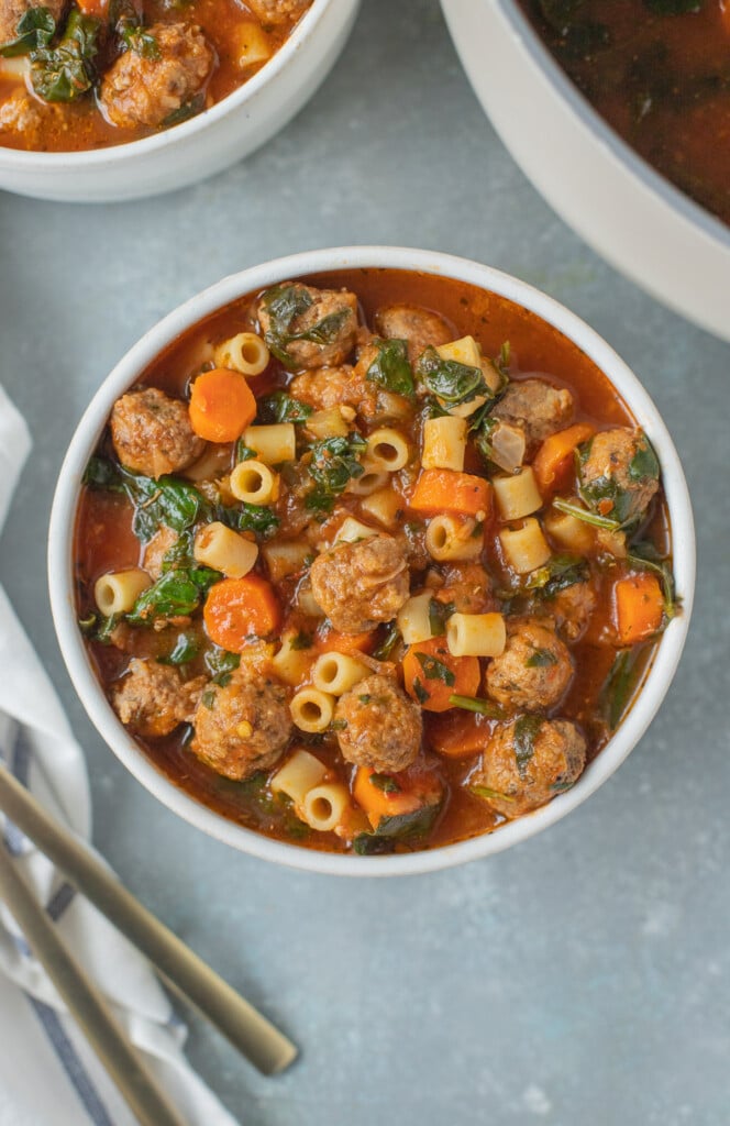 Meatball Soup in a white bowl with a pot and bowl of soup on the side on a grey background