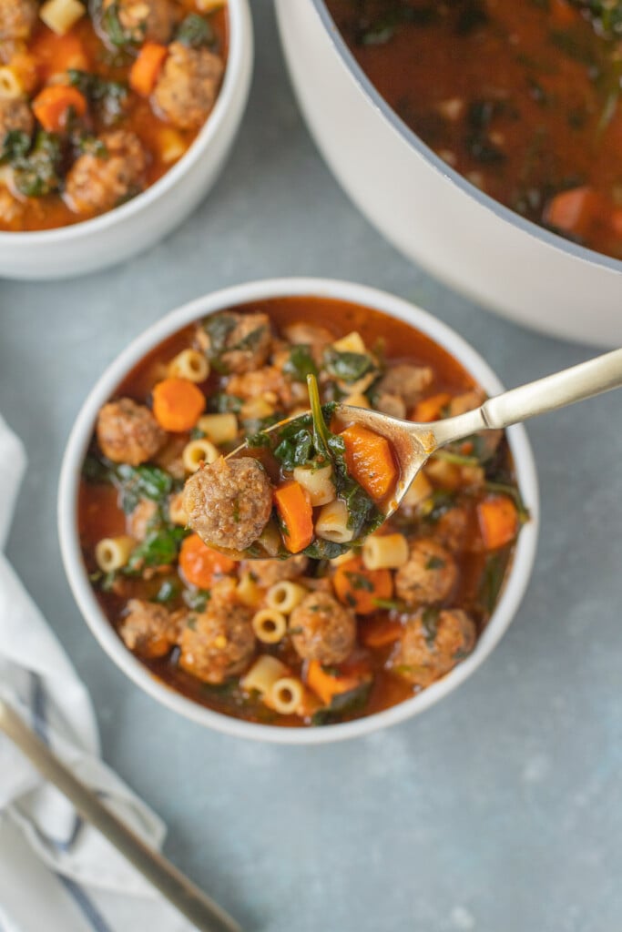 Meatball Soup in a white bowl with a spoon taking a bite out of it with a pot and bowl of soup on the side on a grey background