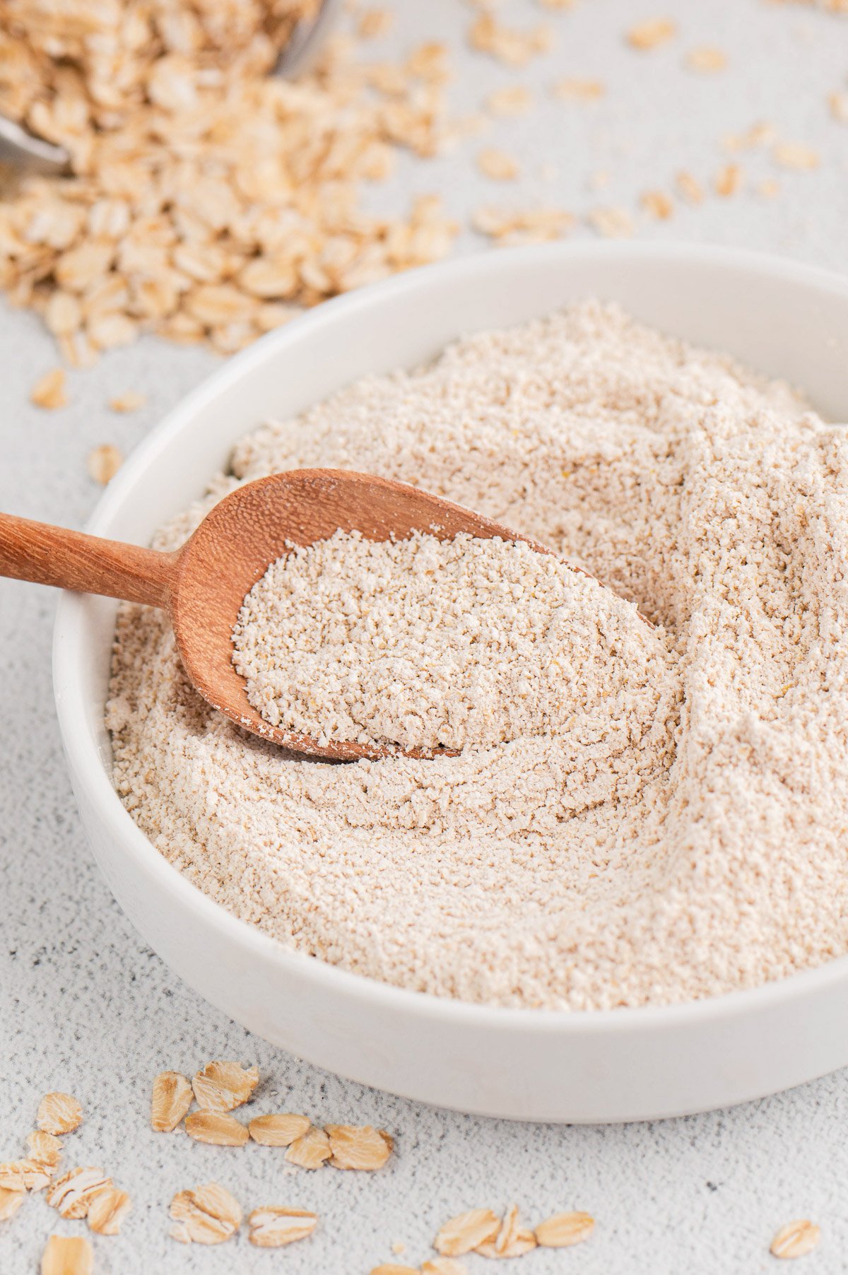 A white bowl of oat flour with a wooden scoop in it with oats in the background