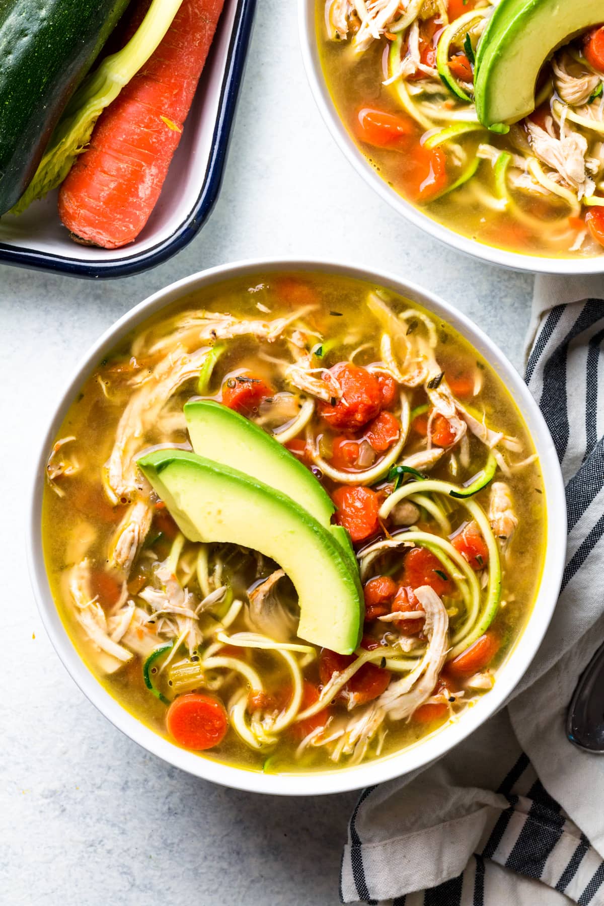A bowl of Paleo Chicken Soup on a grey background with veggies and a linen off to the side