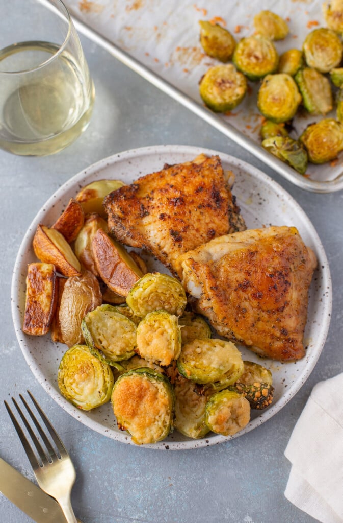 An overhead shot of a plate with parmesan brussel sprouts, roasted fingerling potatoes, and baked chicken thighs with a sheet pan of brussels, a glass of wine, and gold silverware on the side