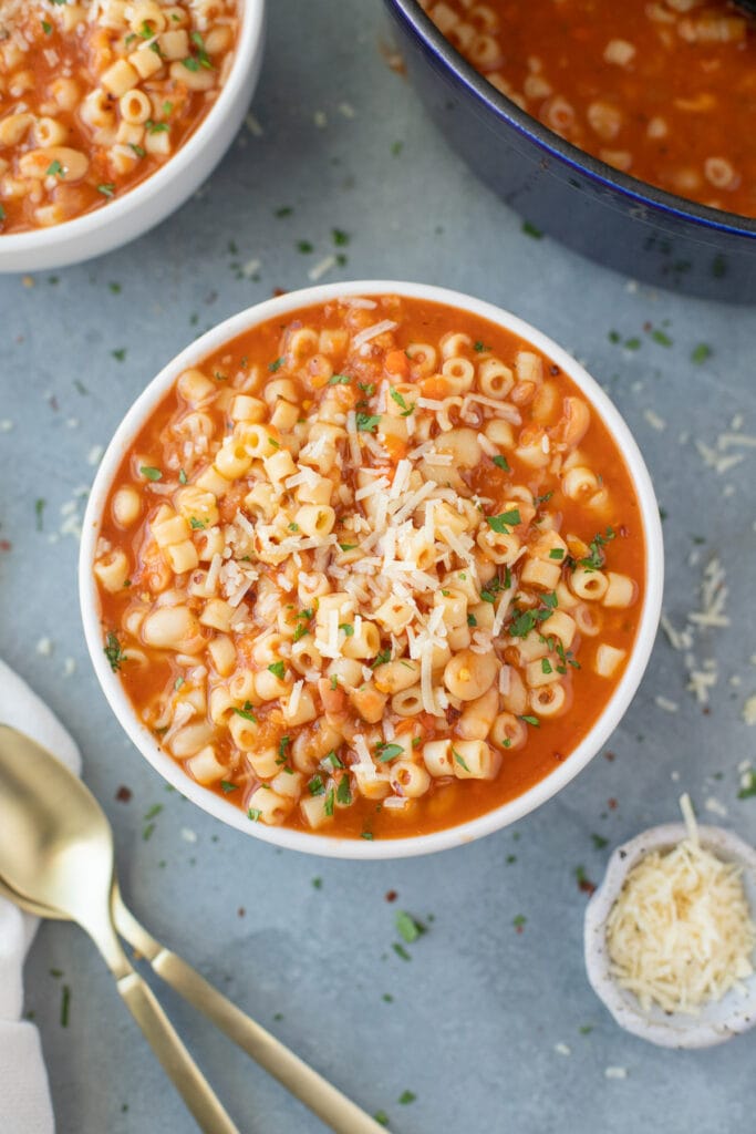 An overhead shot of a bowl of Pasta Fagioli with a bowl and pot of pasta fagioli soup to the side