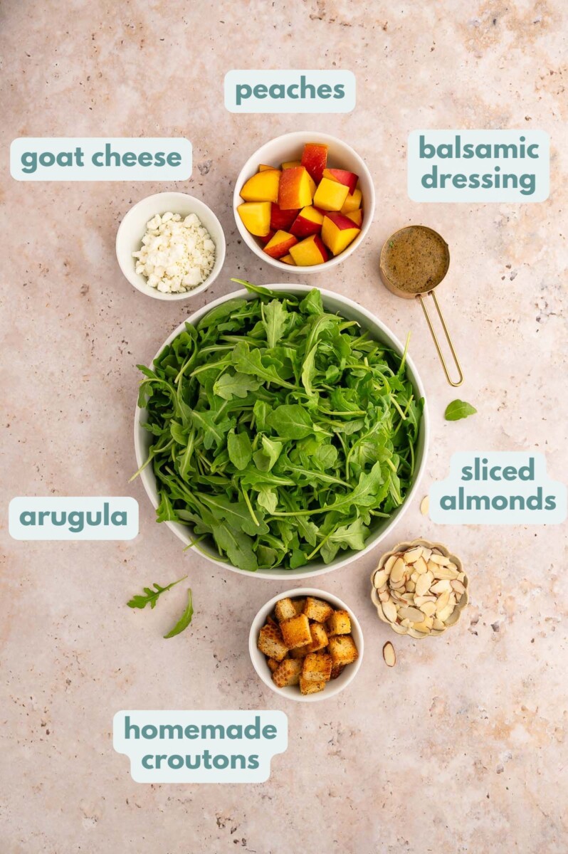 Ingredients for healthy peach salad in small bows: goat cheese, peaches, balsamic dressing, sliced almonds, homemade croutons, arugula